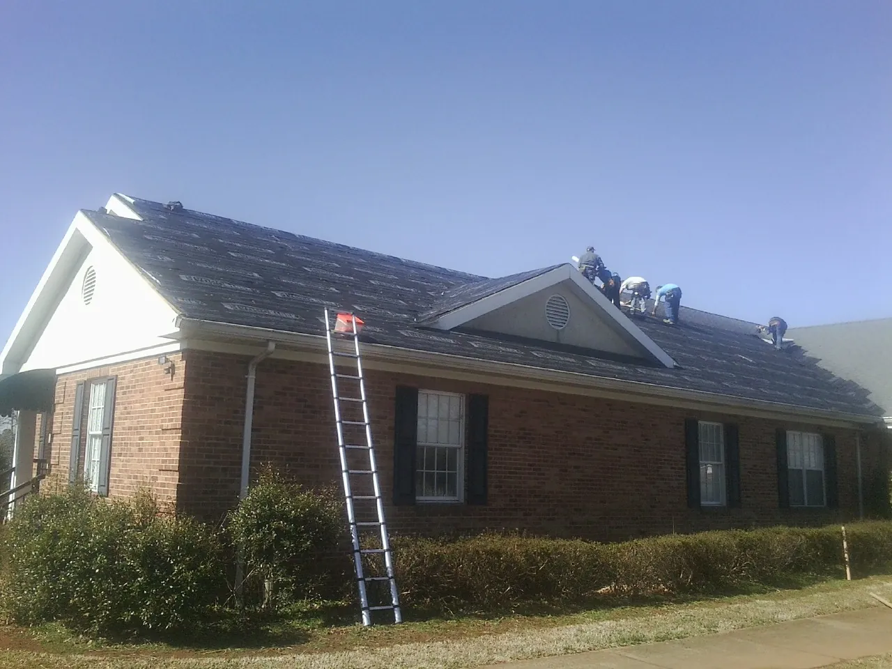 Crew tearing shingles off a brick ranch roof with a ladder set — Emergency Roof Repair work by our crew in Fort Pierre