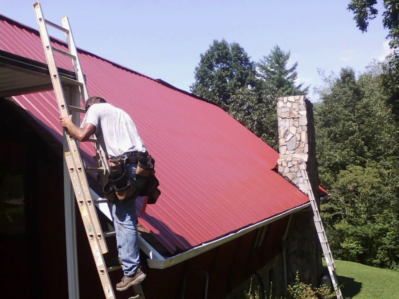 Roofer on a ladder beside a red metal roof and stone chimney — Soffit & Fascia Repair by Roofing Brothers in Fort Pierre